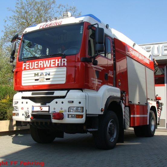 Ein großer roter und weißer Feuerwehrwagen mit der Aufschrift Feuerwehr an der Seite und MAN vorne steht vor einem Feuerwehrgebäude.