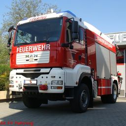 Ein großer roter und weißer Feuerwehrwagen mit der Aufschrift Feuerwehr an der Seite und MAN vorne steht vor einem Feuerwehrgebäude.