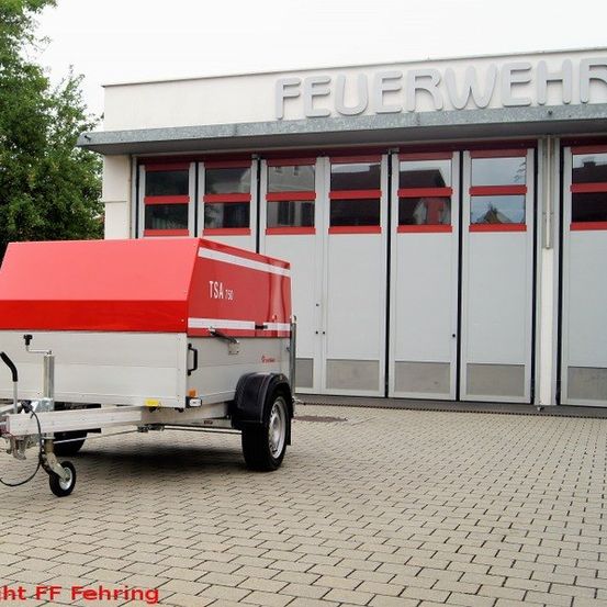 Ein roter und weißer Anhänger steht vor einem Gebäude mit der Aufschrift 'Feuerwehr'.