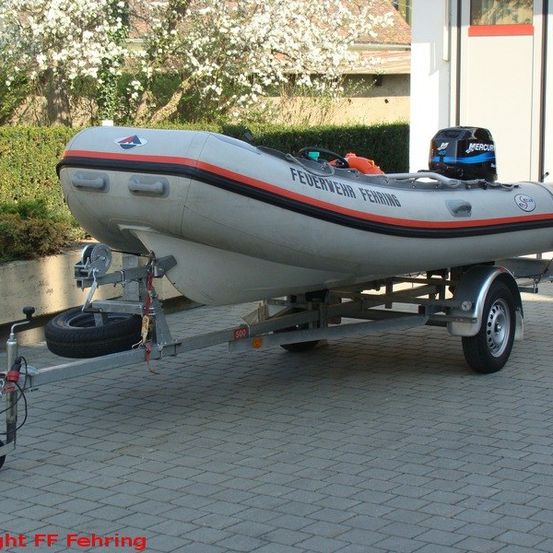 Ein aufblasbares Boot mit schwarzem Motor und einem Reifen steht auf einem Anhänger vor einem Gebäude mit blühenden Bäumen.