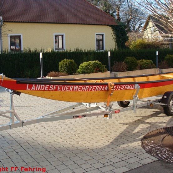 Ein gelbes und rotes Boot namens Landesfeuerwehrverband Steyrmann steht auf einem Anhänger mit einem Haus und einem Baum im Hintergrund.