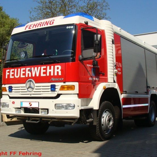 Ein roter und weißer Feuerwehrwagen mit dem Wort 'Feuerwehr' auf der Front ist vor einem Gebäude geparkt. Es ist ein Mercedes-Benz Atego mit Kennzeichen und einer blauen Licht auf der Oberseite.