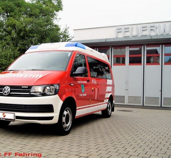 Ein rot-weißer Volkswagen-Bus ist vor einem Gebäude mit der Aufschrift 'Feuerwehr' geparkt.