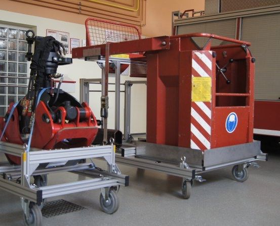 Zwei rote Ausrüstungswagen, jeder mit einem großen mechanischen Gerät, stehen in einer Garage. Dahinter ist ein Feuerwehrauto mit einer roten Tür zu sehen.