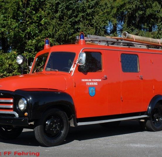 Ein alter roter Feuerwehrwagen ist auf einem Parkplatz abgestellt, mit Bäumen im Hintergrund. Das Nummernschild zeigt 'S9.796'.