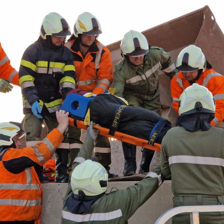 Rettungskräfte tragen eine Person auf einer Trage vor einer Baustelle. Sie tragen alle Schutzausrüstung.