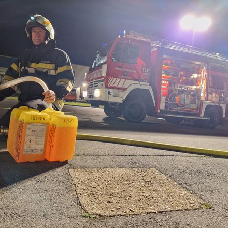 Ein Feuerwehrmann hält einen Schlauch an orangefarbenen Tanks. Ein roter Feuerwehrwagen mit ausgefahrener Leiter steht im Hintergrund.