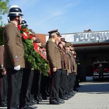 Uniformierte Feuerwehrleute stehen mit einem Kranz in einer Reihe vor der Feuerwache. Ein Krankenwagen ist in der Station geparkt.