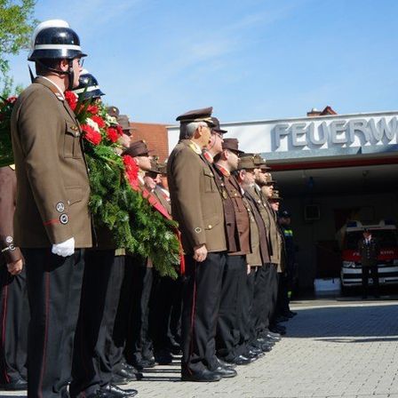 Uniformierte Feuerwehrleute stehen mit einem Kranz in einer Reihe vor der Feuerwache. Ein Krankenwagen ist in der Station geparkt.