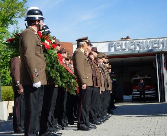 Uniformierte Feuerwehrleute stehen mit einem Kranz in einer Reihe vor der Feuerwache. Ein Krankenwagen ist in der Station geparkt.