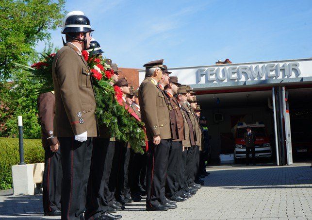 Uniformierte Feuerwehrleute stehen mit einem Kranz in einer Reihe vor der Feuerwache. Ein Krankenwagen ist in der Station geparkt.