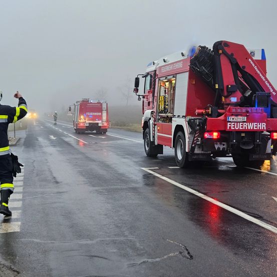 Feuerwehrleute leiten den Verkehr auf einer nebligen Straße, mit einem Feuerwehrauto im Hintergrund. Ein Feuerwehrmann steht an der Seite und signalisiert den Fahrzeugen.