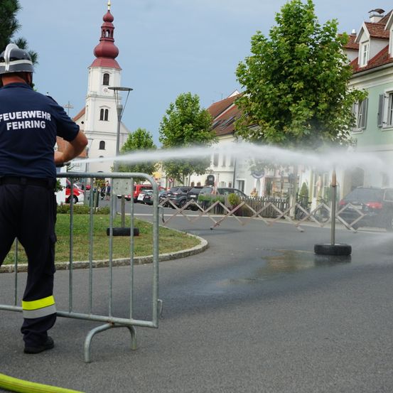 Ein Feuerwehrmann sprüht Wasser aus einem Schlauch auf die Straße und steht hinter einem Metallzaun. Die Szene spielt sich auf einer Straße ab, die von Bäumen und geparkten Autos gesäumt ist. Im Hintergrund befindet sich eine Kirche mit einem roten Turm.