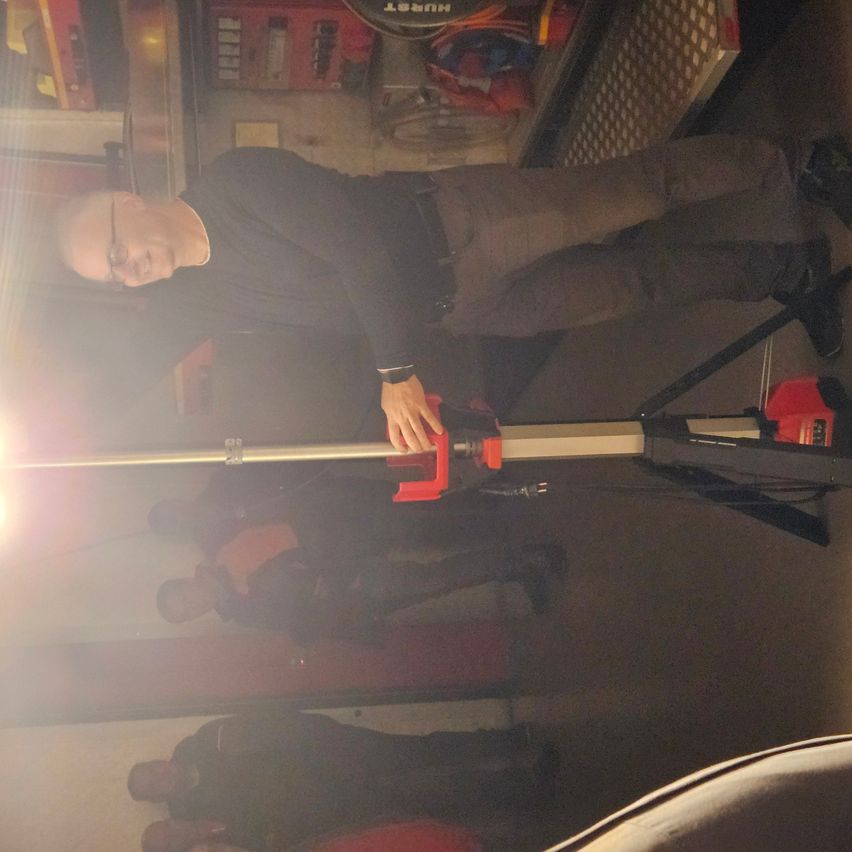 A man with glasses is holding a lit flashlight, possibly a demonstration, with a crowd watching. The flashlight is mounted on a tripod.