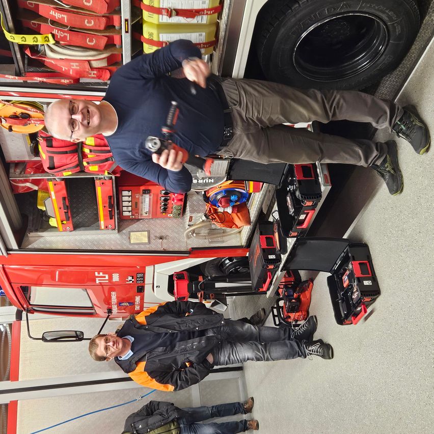 Two men inside a fire truck, one standing holding a tool, the other sitting, both smiling. The fire truck has tools and equipment.