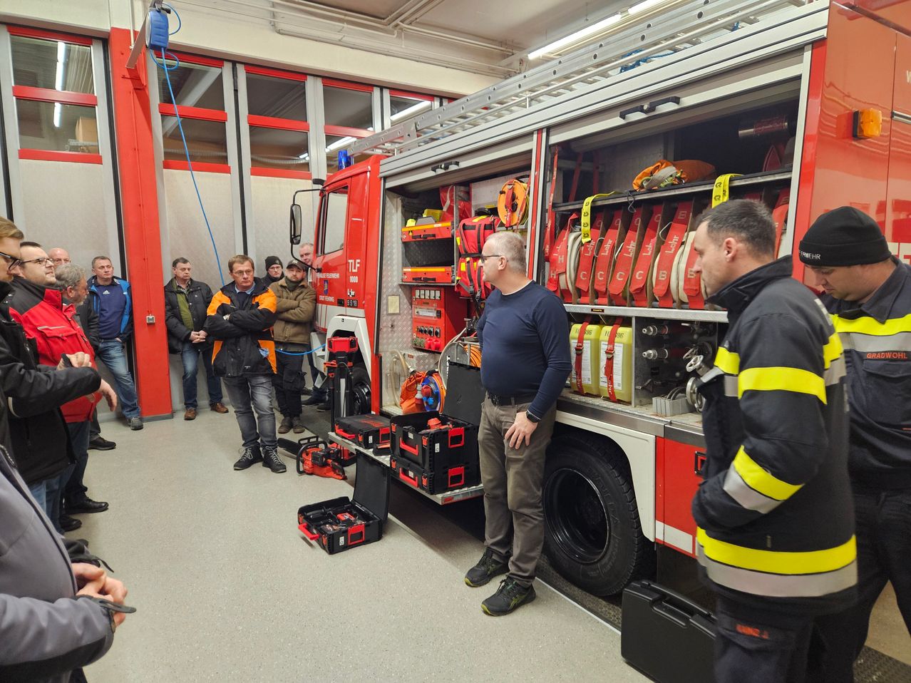 Inside a fire station, a group of men stand around a fire truck. A man with glasses and a watch speaks to others. Various equipment and tools are visible on the truck and floor.