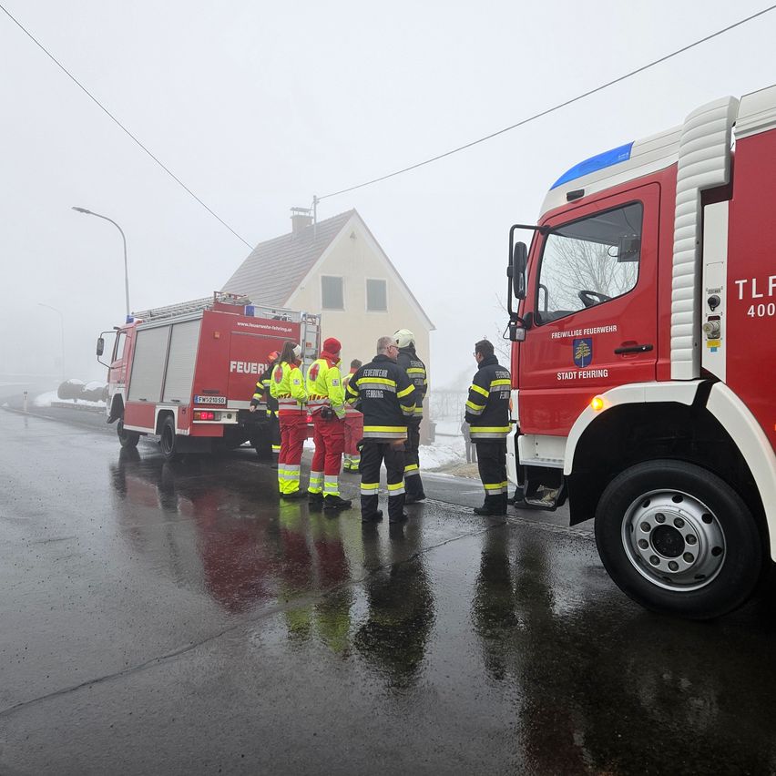 Firefighters in reflective gear stand by a red fire truck on a wet road with fog in the background.