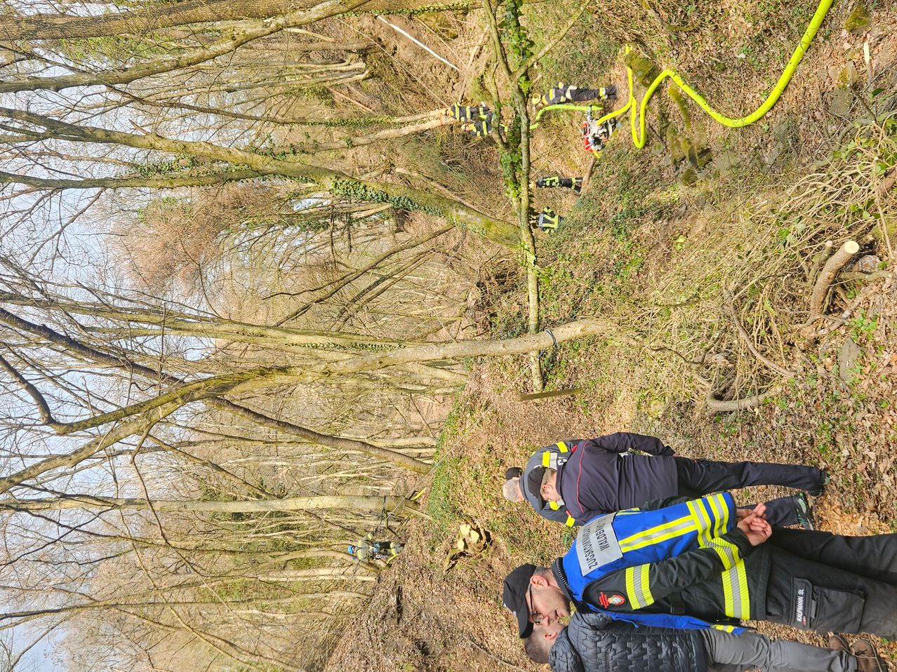 Rettungskräfte in reflektierenden Westen arbeiten an einem Hang mit einer gelben Schlange, umgeben von kahlen Bäumen.