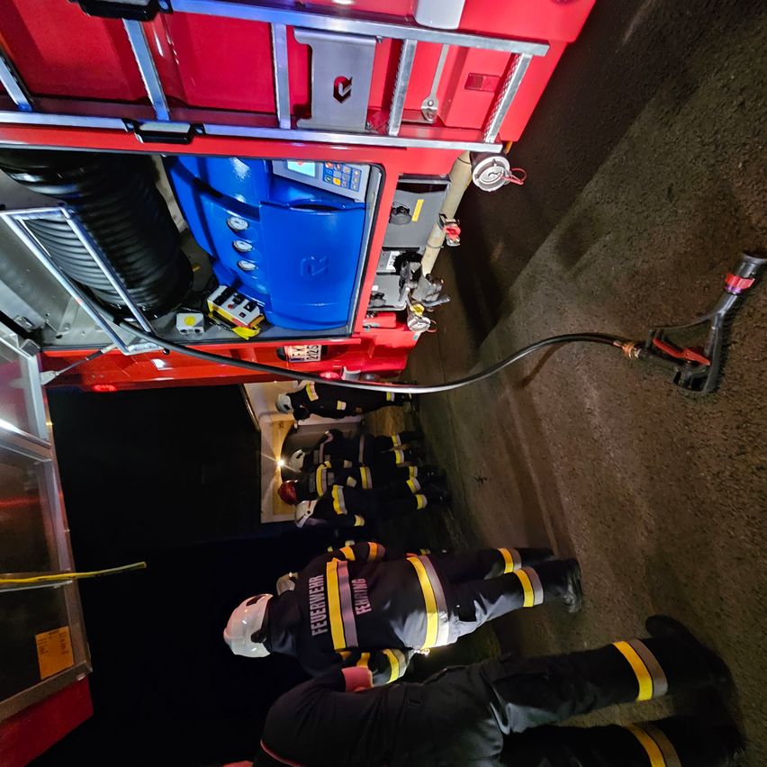 Feuerwehrleute in Schutzkleidung sind in einem Fahrzeug und bereiten sich auf einen Notfall vor. Ein Schlauch ist angeschlossen, und die Ausrüstung des Fahrzeugs ist sichtbar.
