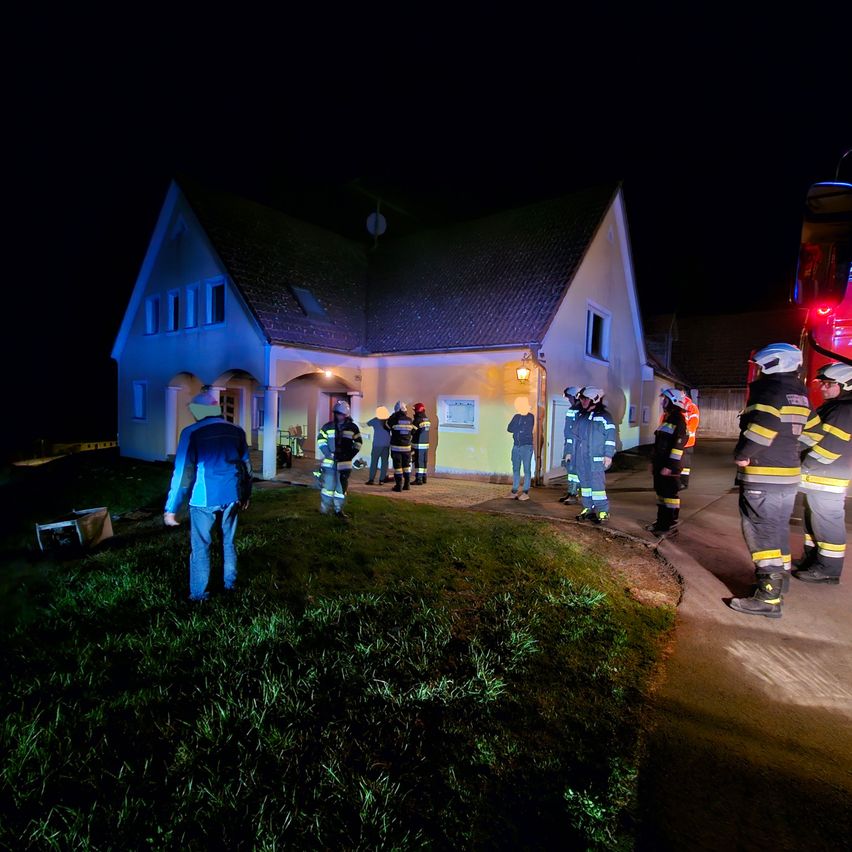 Eine Gruppe Feuerwehrleute in voller Ausrüstung steht nachts vor einem Haus. Eine Person in einem blauen Mantel steht in der Nähe des Eingangs.