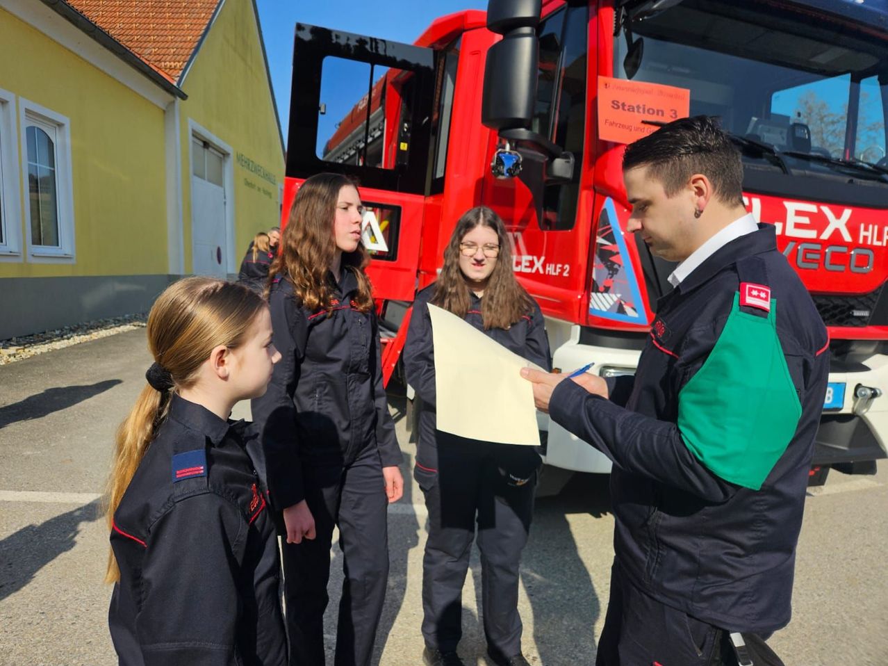 Fünf Personen in Feuerwehruniformen, darunter drei Frauen und ein Mann, stehen vor einem roten Feuerwehrauto mit der Aufschrift Station 3. Ein Mann in Uniform hält ein Stück Papier und einen Stift.