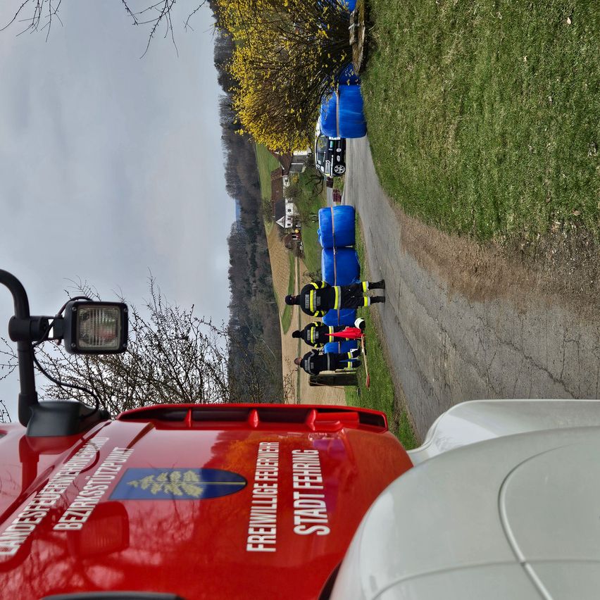 Ein rotes Fahrzeug mit Notfalltext auf der Rückseite. Eine Straße mit blauen Fässern und Personen in Sicherheitskleidung an der Seite.