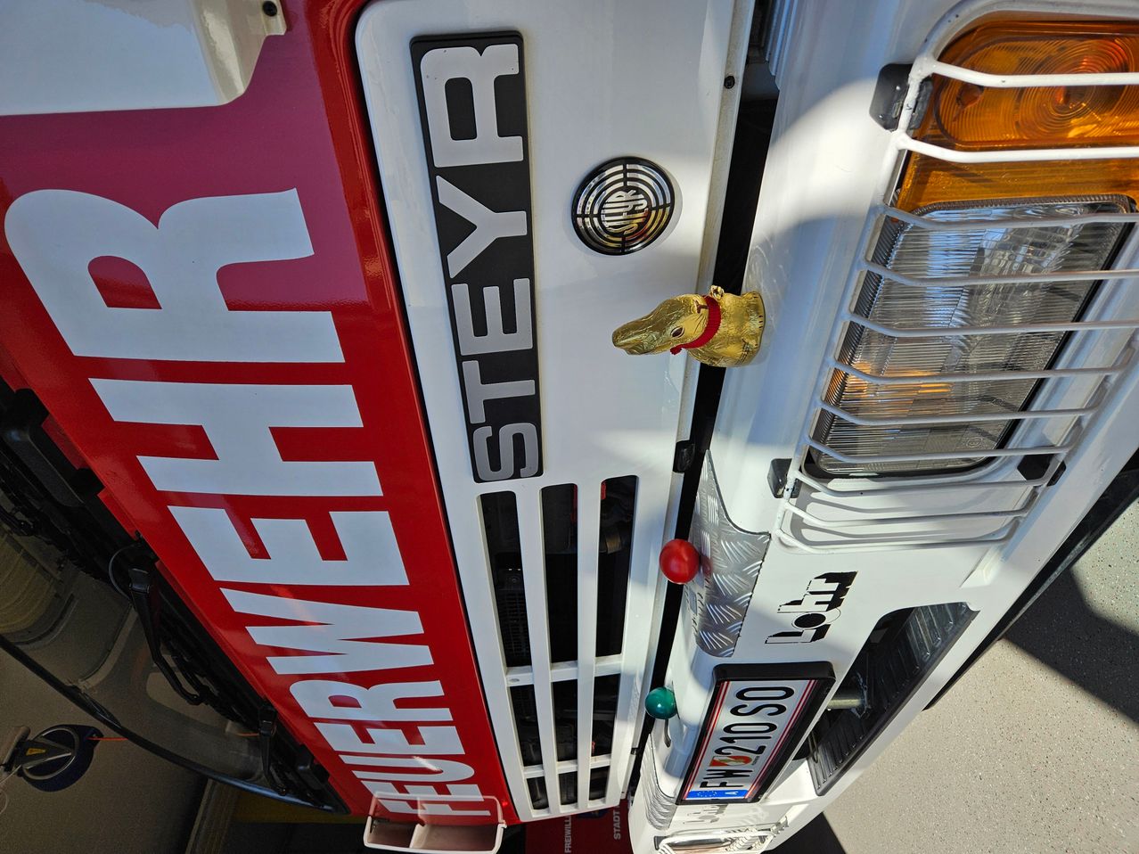 Eine Nahaufnahme eines weißen und roten Fahrzeugs mit dem Wort 'FEUERWEHR' in Rot geschrieben. Es gibt eine goldene Hundefigur vorne und ein Kennzeichen, das 'FW 210 SO' liest.