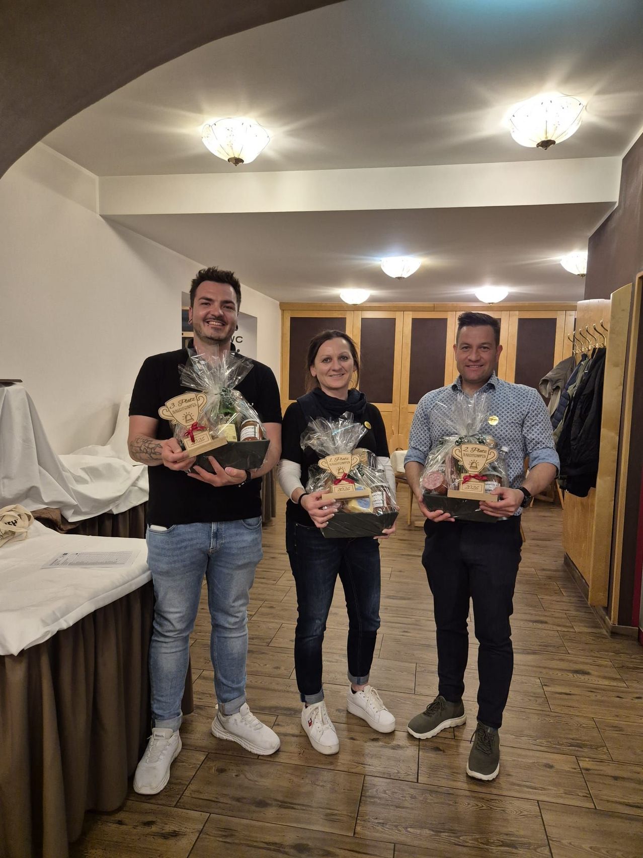 Drei Menschen stehen in einem Raum mit Holzböden. Sie halten Geschenkkörbe und lächeln. Der Raum hat Holzschränke und einen Tisch, der mit einem weißen Tuch bedeckt ist.