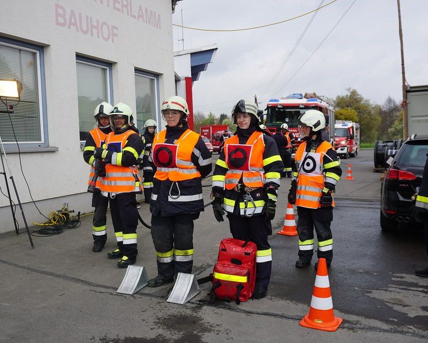 Fünf Feuerwehrleute stehen vor einem Gebäude mit einem roten Schild, das 'Bauhof' besagt. Sie tragen reflektierende Westen und Helme. In der Nähe gibt es einen roten Rucksack und orangefarbene Verkehrskegel. Hinter ihnen sind Feuerwehrfahrzeuge geparkt.