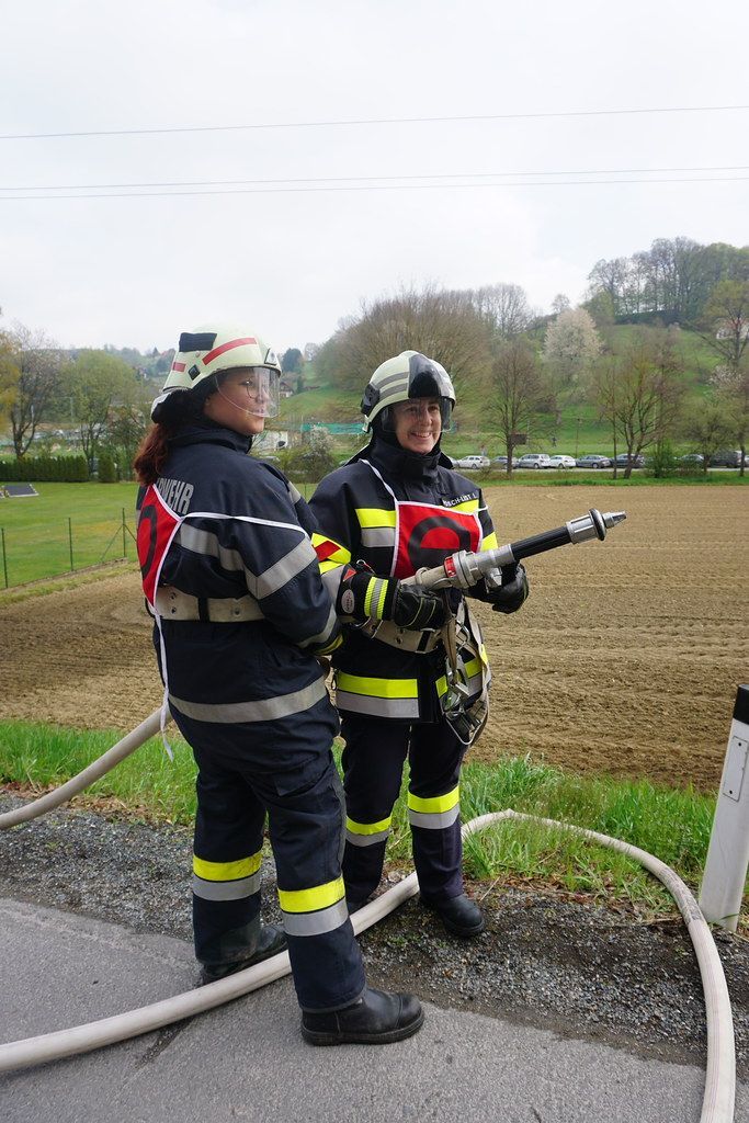 Zwei Feuerwehrfrauen stehen neben einem Feuerlöscher, eine hält eine Schlauch. Sie tragen Uniformen mit reflektierenden Streifen. Dahinter erstreckt sich ein Feld mit spärlicher Vegetation und geparkten Autos.