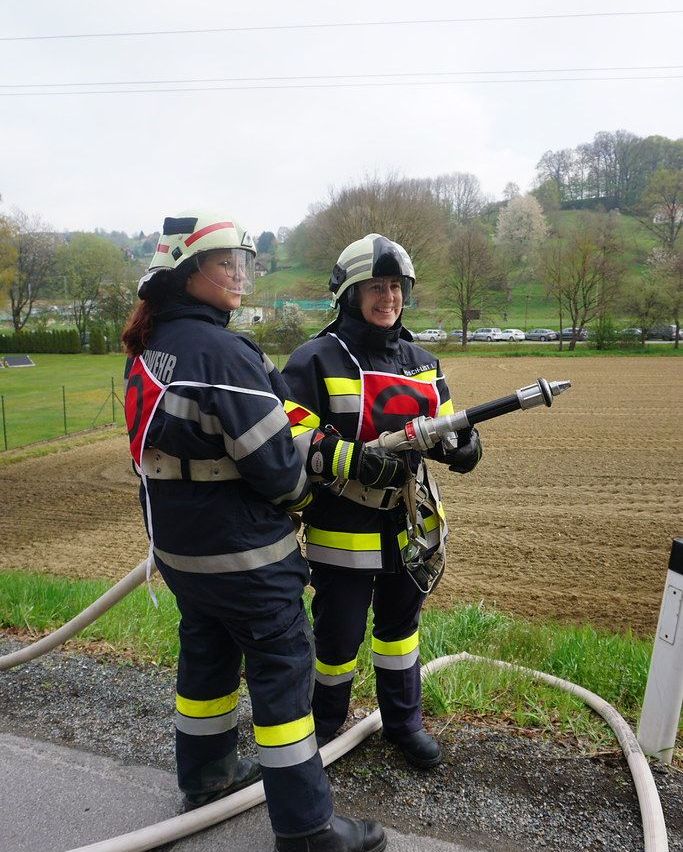 Zwei Feuerwehrfrauen stehen neben einem Feuerlöscher, eine hält eine Schlauch. Sie tragen Uniformen mit reflektierenden Streifen. Dahinter erstreckt sich ein Feld mit spärlicher Vegetation und geparkten Autos.