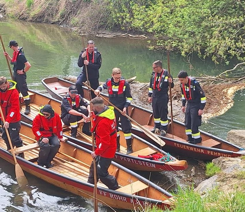 Eine Gruppe Feuerwehrleute in roten Jacken bereitet ihre Kanus am Flussufer vor. Sie halten Paddel und tragen Sicherheitsausrüstung.