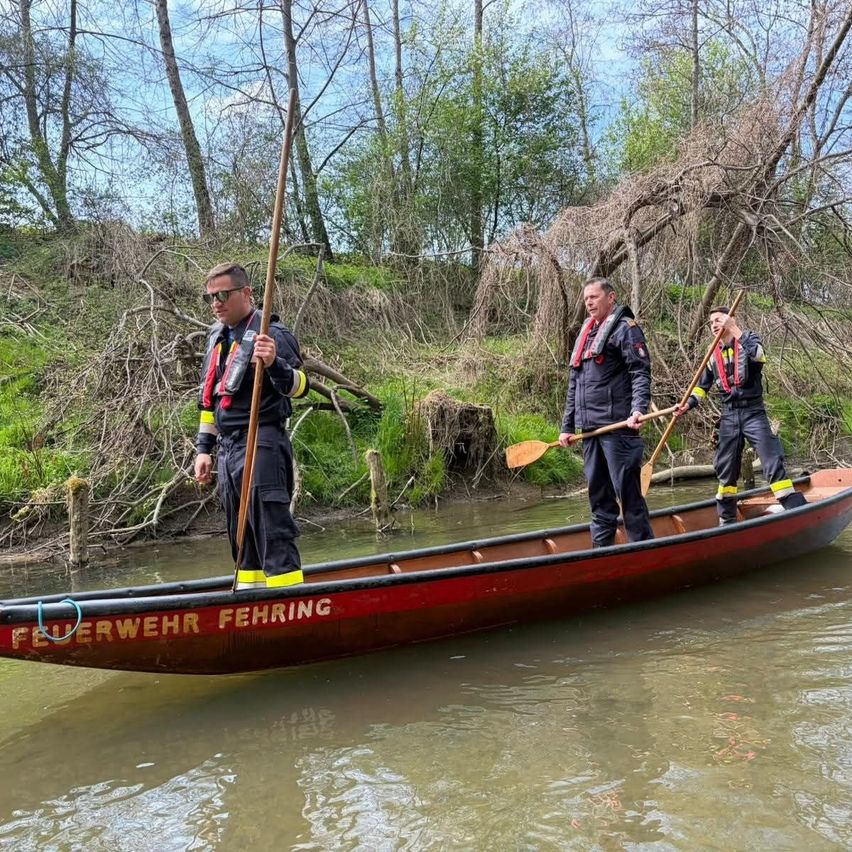 Drei Feuerwehrleute in Sicherheitswesten paddeln ein Kanu auf einem Fluss. Einer steht vorne, die anderen stehen hinter ihm. Das Kanu hat das Wort 'Feuerwehr' darauf geschrieben.
