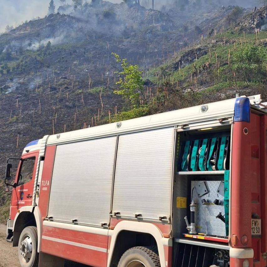 Ein roter Feuerwehrwagen mit offenen Türen steht auf einer unbefestigten Straße. Er hat einen Aufbewahrungskasten im Inneren und ein Kennzeichen mit der Aufschrift FW 12 SO. Im Hintergrund befindet sich ein Hügel mit Bäumen und Pflanzen und Rauch von einem Feuer.