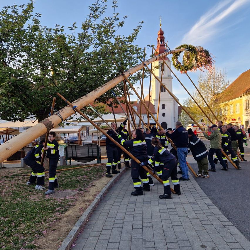 Feuerwehrleute heben einen großen, mit einem Kranz verzierten Holzpfahl an, während sie auf einem gepflasterten Gehweg stehen. Dahinter befindet sich eine Kirche mit einem Turm.