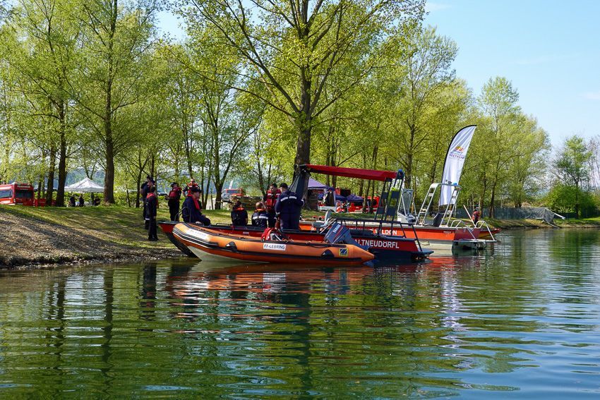 Rettungskräfte in roten Jacken stehen neben Booten am Seeufer, mit Bäumen und Zelten im Hintergrund.
