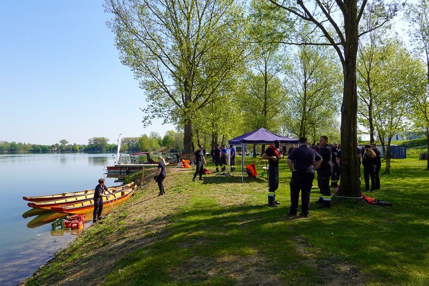 Men in Uniform stehen in der Nähe von Booten und einem Zelt am See. Einige stehen, andere sitzen auf Bänken. Bäume und ein Gebäude sind im Hintergrund.