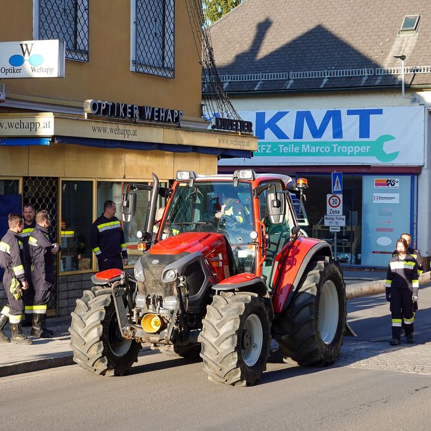 Ein roter Traktor ist am Straßenrand geparkt. Mehrere Feuerwehrleute in Uniformen stehen in der Nähe. Ein Gebäude mit einem großen Schild, das "KMTT" liest, befindet sich im Hintergrund.