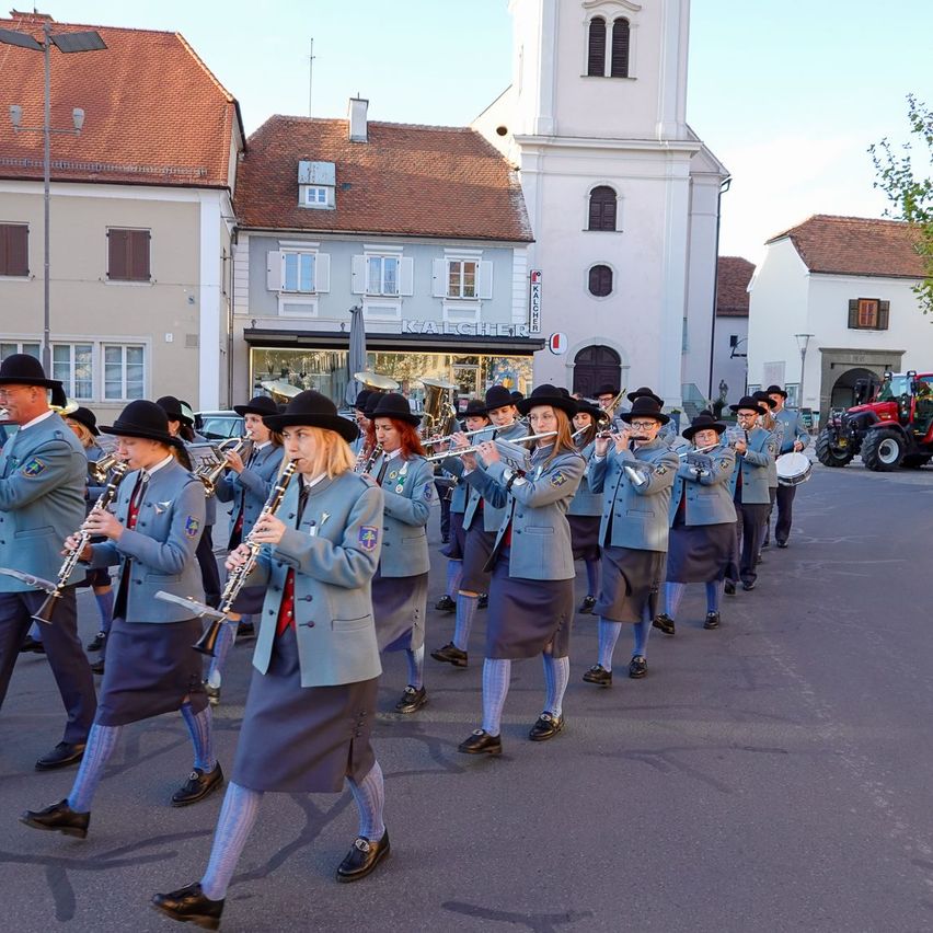 Ein Marschkapelle in einheitlichen Uniformen tritt vor Gebäuden im Freien auf. Einige spielen Flöten, während andere Trompeten spielen. Die Band marschiert auf einer asphaltierten Straße.