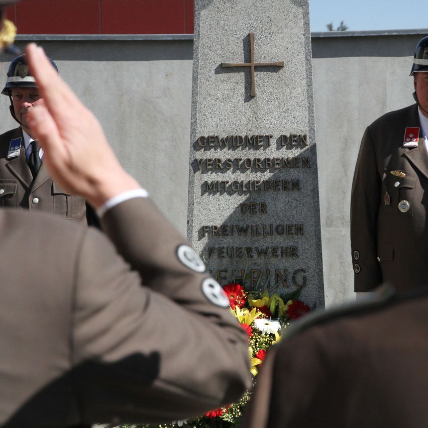 Soldaten salutieren vor einem Denkmal mit einem Kreuz, Blumen und deutschem Text. Ein Blumenkranz liegt vor dem Denkmal.