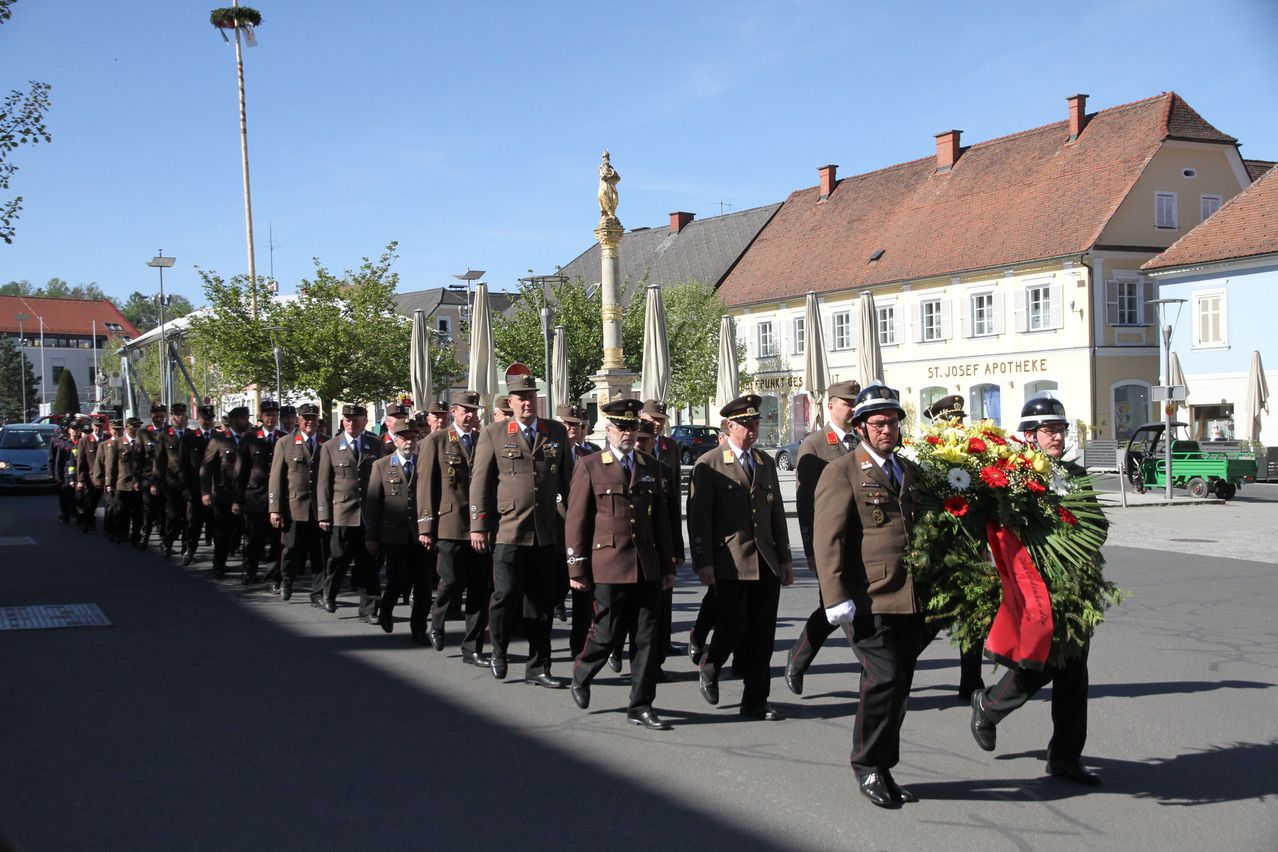 Eine Gruppe von Militärangehörigen in braunen Uniformen marschiert bei einem Paradezug und trägt einen Kranz, unter einem klaren Himmel.