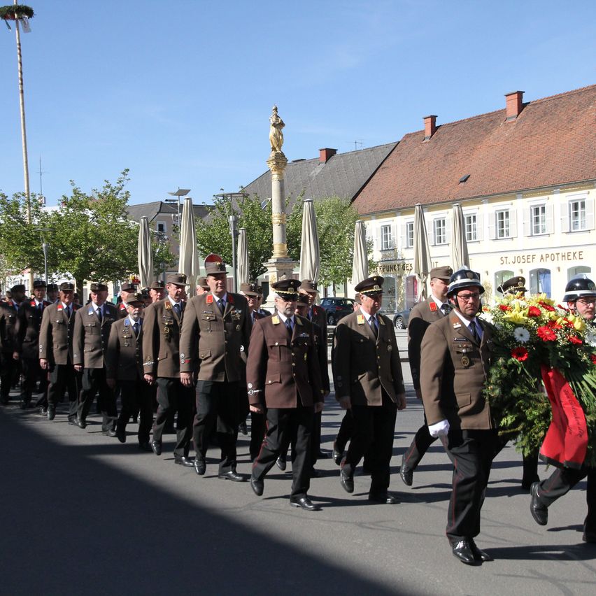Eine Gruppe von Militärangehörigen in braunen Uniformen marschiert bei einem Paradezug und trägt einen Kranz, unter einem klaren Himmel.