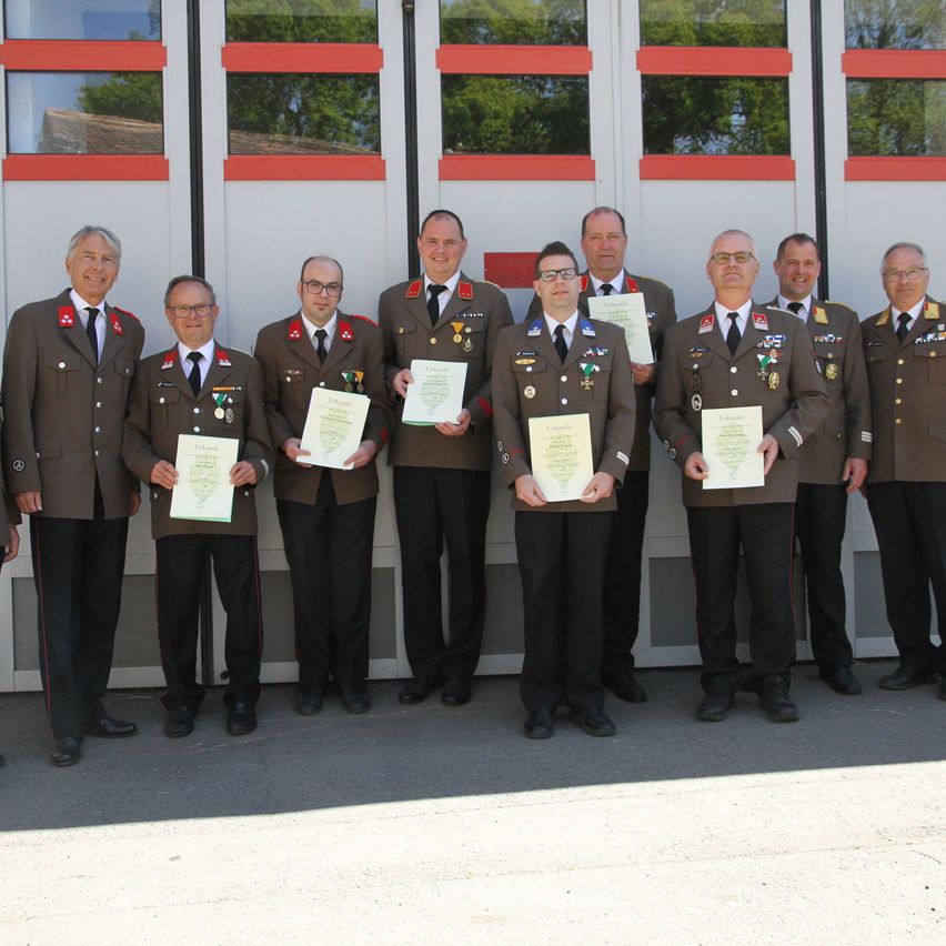 Eine Gruppe von Männern in Uniform steht vor einem Gebäude mit Glasfenstern und hält Urkunden. Sie tragen Medaillen und schwarze Anzüge. Sie scheinen ein formelles Ereignis zu feiern.