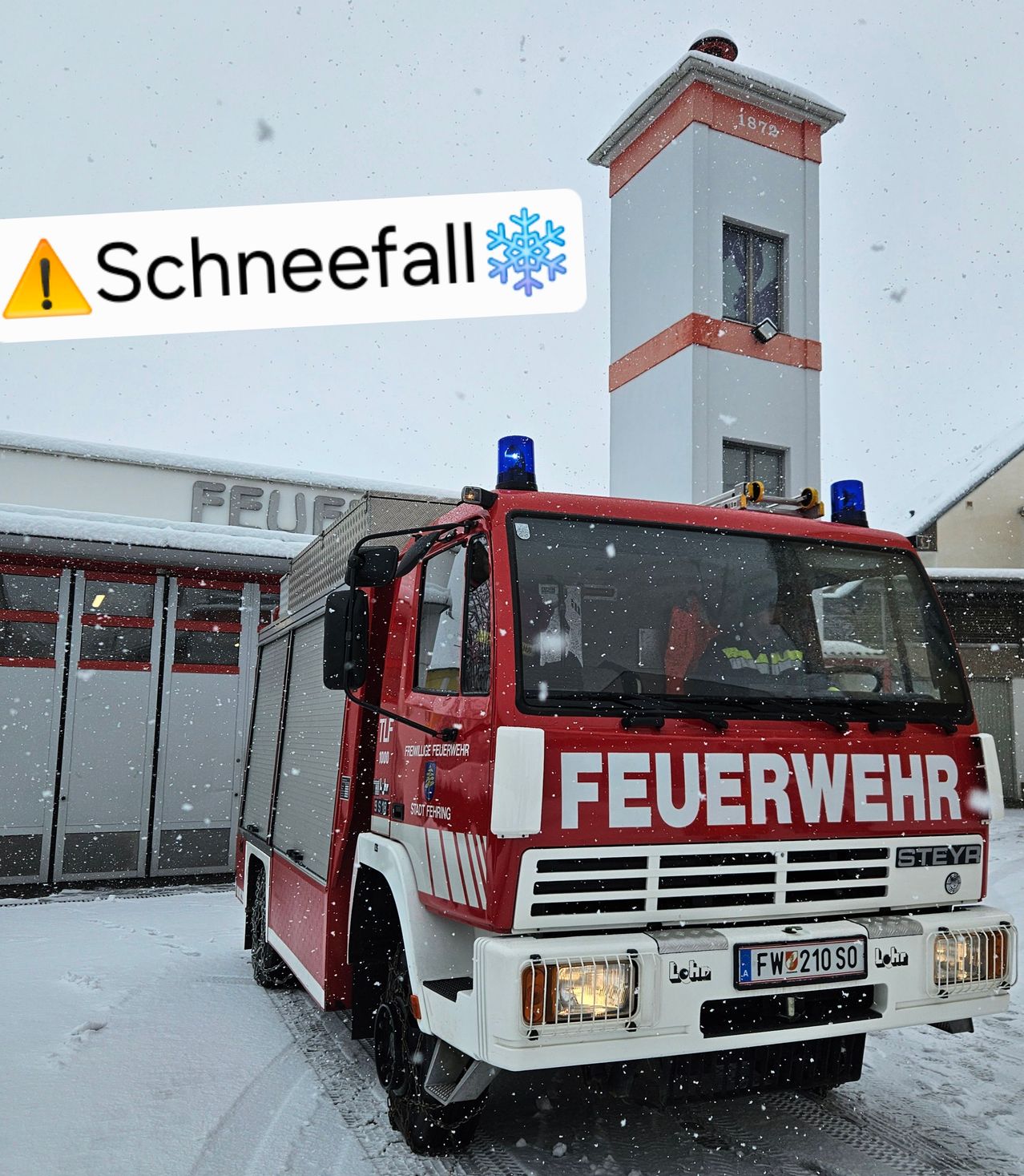 Ein Feuerwehrauto mit der Aufschrift 'Feuerwehr' steht im Schnee vor einem Gebäude mit einem Turm. Über dem Auto ist ein Warnschild für Schneefall zu sehen.