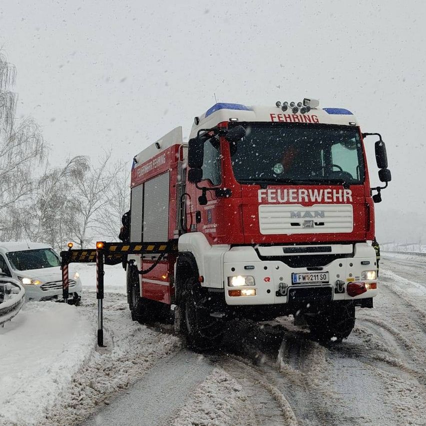 Ein rot-weißer Feuerwehrwagen fährt auf einer verschneiten Straße, in der Nähe ist ein Van im Schnee steckengeblieben.