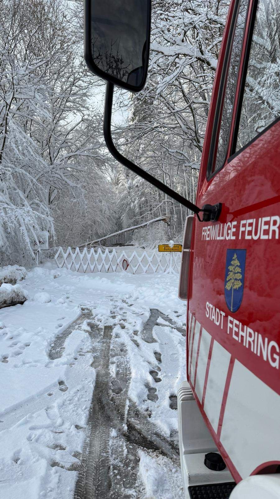 Ein Feuerwehrfahrzeug steht auf einer verschneiten Straße mit schneebedeckten Bäumen. An der Seite des Fahrzeugs steht 'Freiwillige Feuer' und 'Stadt Fehring.' Hinter dem Fahrzeug befindet sich ein weißer Zaun und ein gelbes Schild.