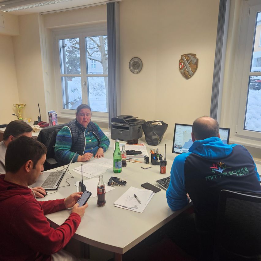 Eine Gruppe von Menschen sitzt um einen Tisch in einem Raum mit verschneiten Fenstern. Sie halten eine Besprechung, mit Laptops und Papieren auf dem Tisch. Ein Pokal steht auf einem Regal.