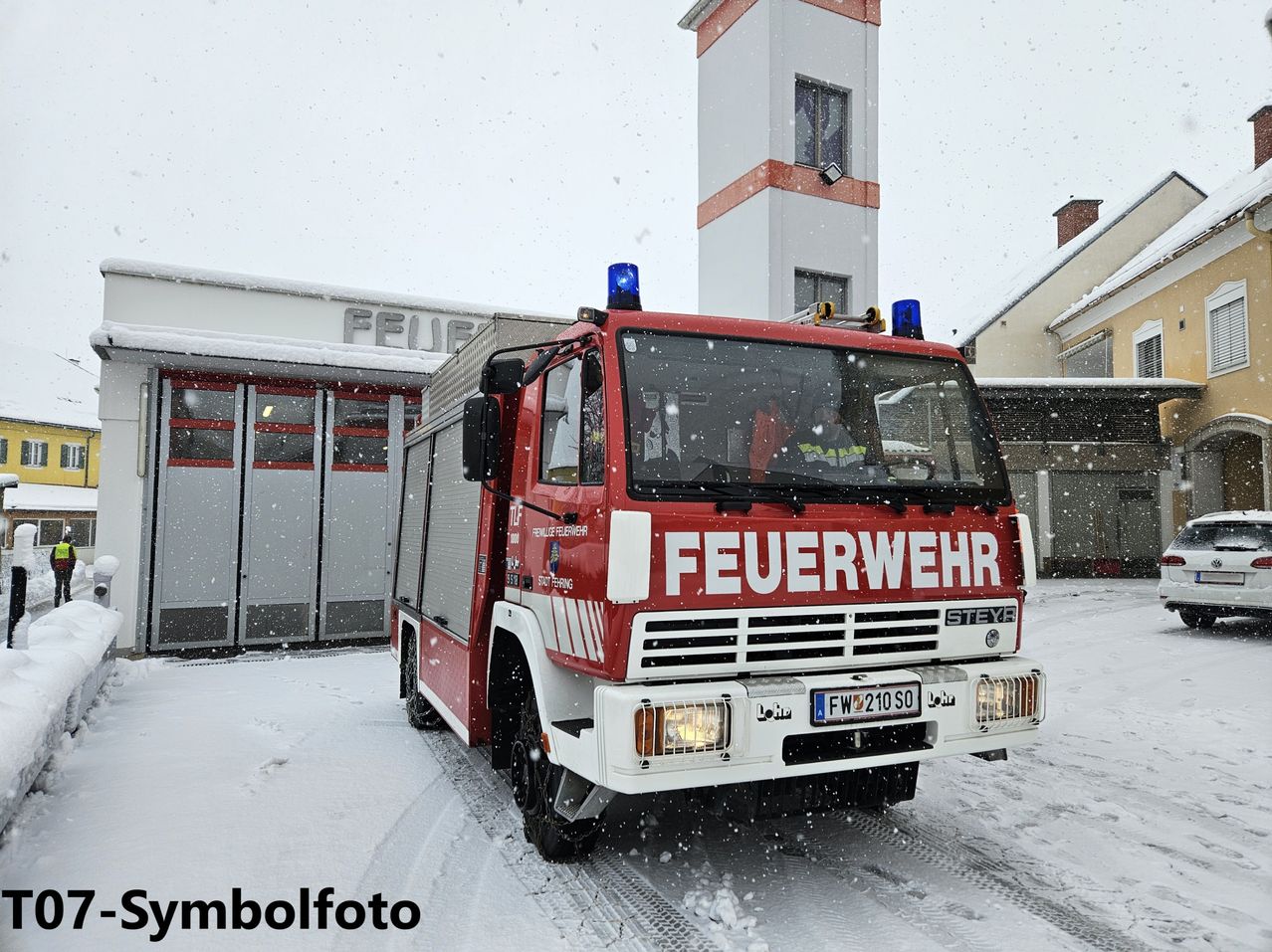 Ein roter Feuerwehrwagen steht auf einer verschneiten Straße mit einem Turm und Gebäude im Hintergrund.