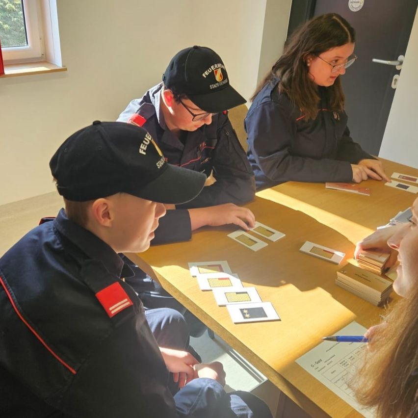 Eine Gruppe von Menschen in Uniformen sitzt um einen Tisch. Sie schauen sich ein Kartenspiel an. Eine der Personen hält einen Stift und schreibt auf ein Papier.