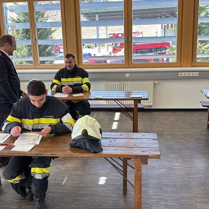 Drei Feuerwehrleute sitzen an einem Tisch in einem Raum und studieren ein Dokument. Ein weiterer Mann steht hinter ihnen. Es gibt andere Tische und ein Feuerwehrhelm liegt auf einem davon. Draußen sind Rettungsfahrzeuge und Bäume zu sehen.